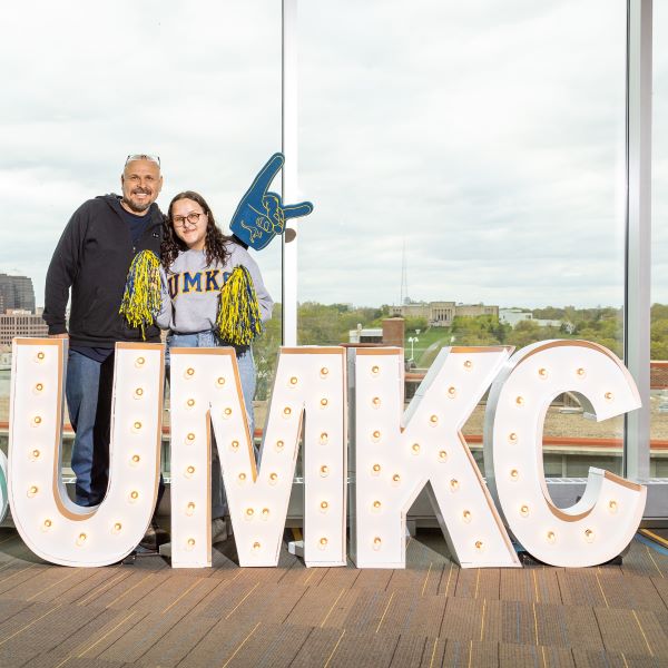 a student holding blue and gold pom poms stands by her father who's holding a UMKC Roos foam finger in front of a UMKC lighted sign on the UMKC Student Union rooftop