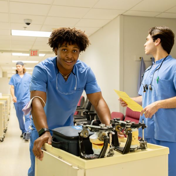 A dental student pushes a cart with equipment down a hall. Another student assists him. They are both wearing blue scrubs.