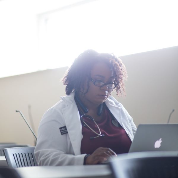 a woman sits at a table with a laptop open. She wears a white health studies lab coat.