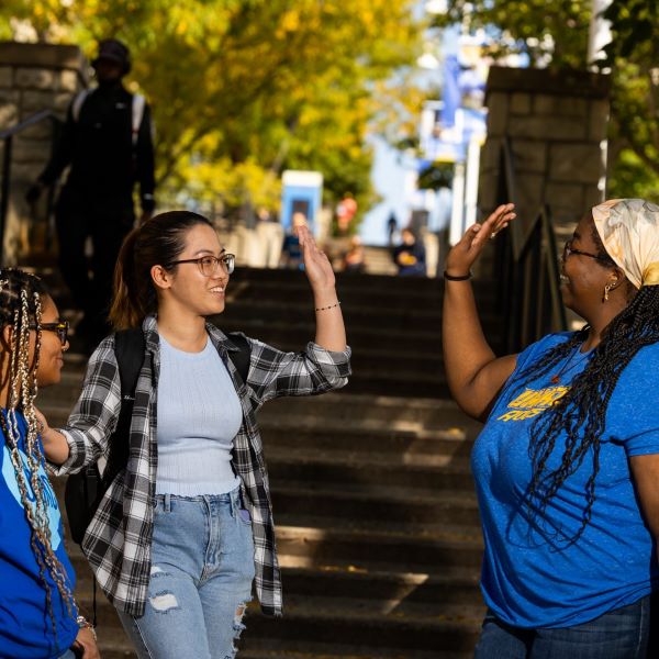 three students stand at the bottom of the UMKC stairs leading up to the Student Union. Two of the students are wearing blue UMKC shirts. One of these students who is wearing a scarf around her braids is giving another student a high five.