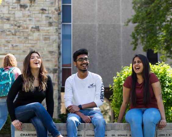 three students sit on a bench outside on UMKC campus. They are laughing.