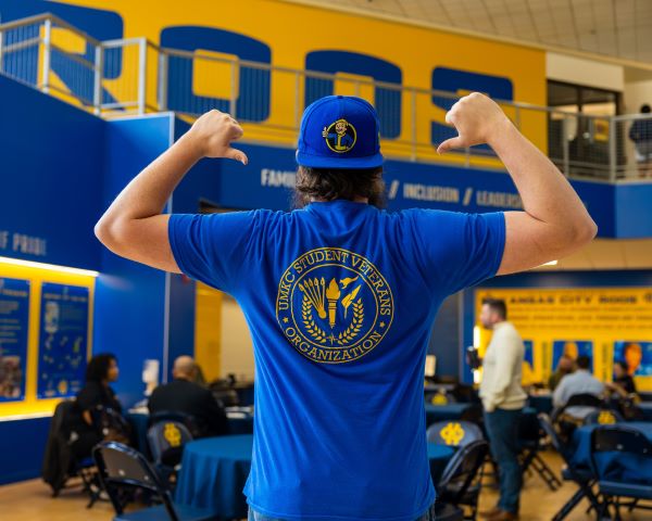 a student has his back to the camera and points to a UMKC Student Veterans logo on a blue t-shirt