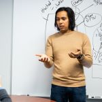 Riddhiman Das concentrates as he turns from a whiteboard to talk to someone sitting at a table off camera. He wears a tan sweater.