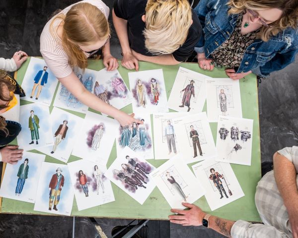 Students in the costume shop gather around a table wand point at beautiful and detailed sketches of costumes for a production.