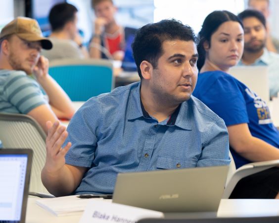 A student sitting at a table in front of an open laptop makes a comment in a class at the Bloch School. He is wearing a blue button-down shirt. Other students sit behind and beside him.