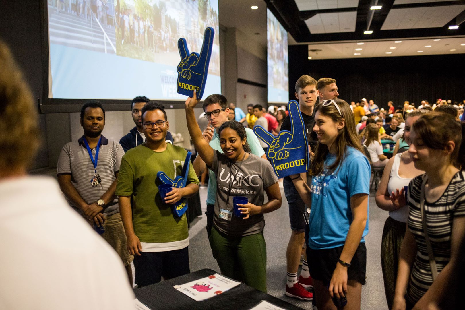 Students holding Roo Up foam fingers gather at a table in an auditorium while they wait for Orientation to begin