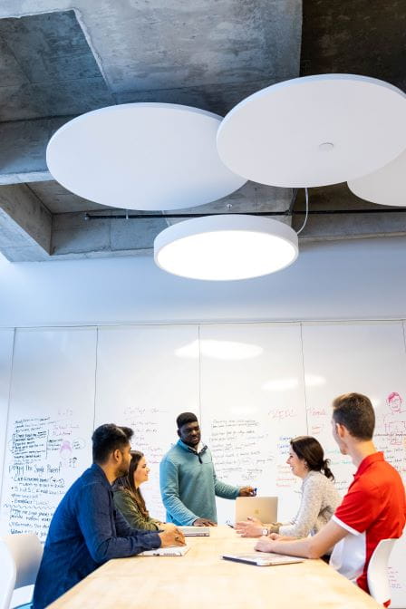 group of students and a professor sit at a round white table in front of a white board pinned with papers in neat rows