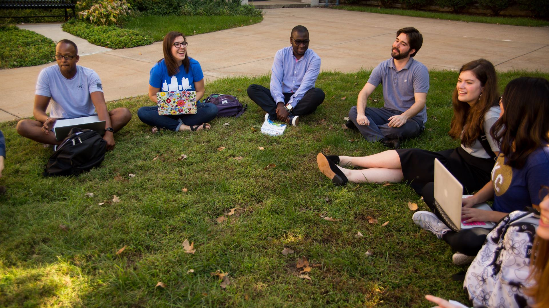 students sit in a circle on the grass on the campus lawn. They are smiling and laughing while having a discussion with their political science professor. Some have open laptops in their laps.