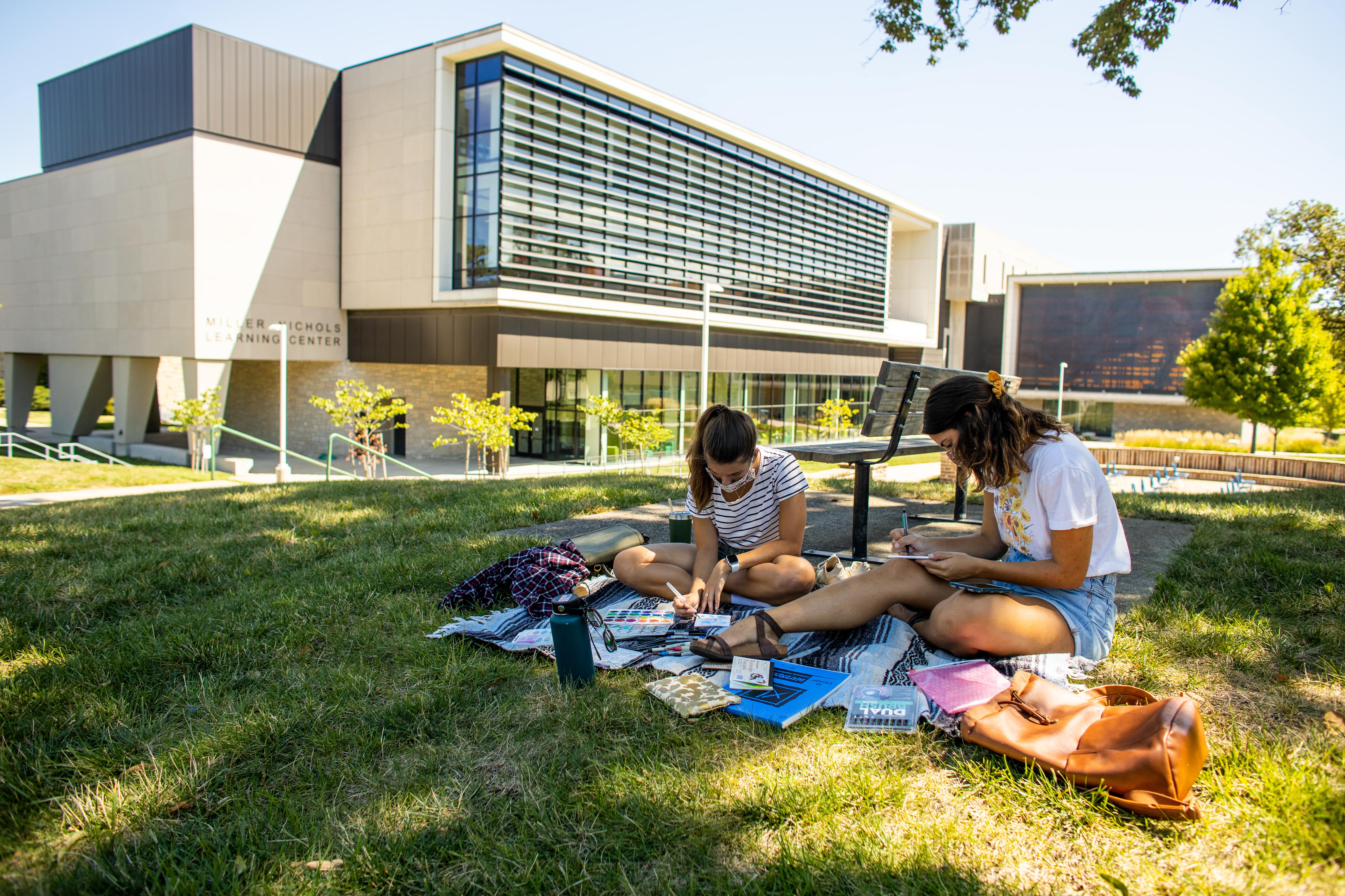 students by the library, painting on a blanket outside on a sunny day.