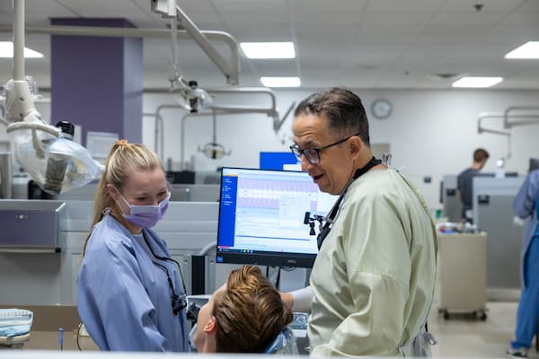 dental student and professor stand in dental lab while patient sits in chair