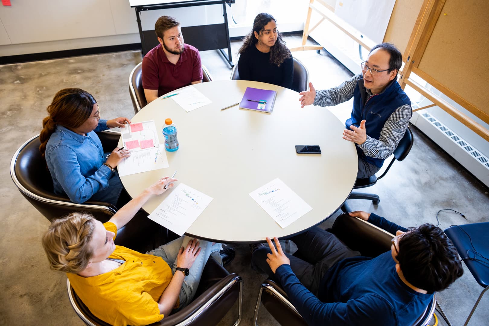 a professor holds his arms in the air while discussing a topic with students sitting around a round table