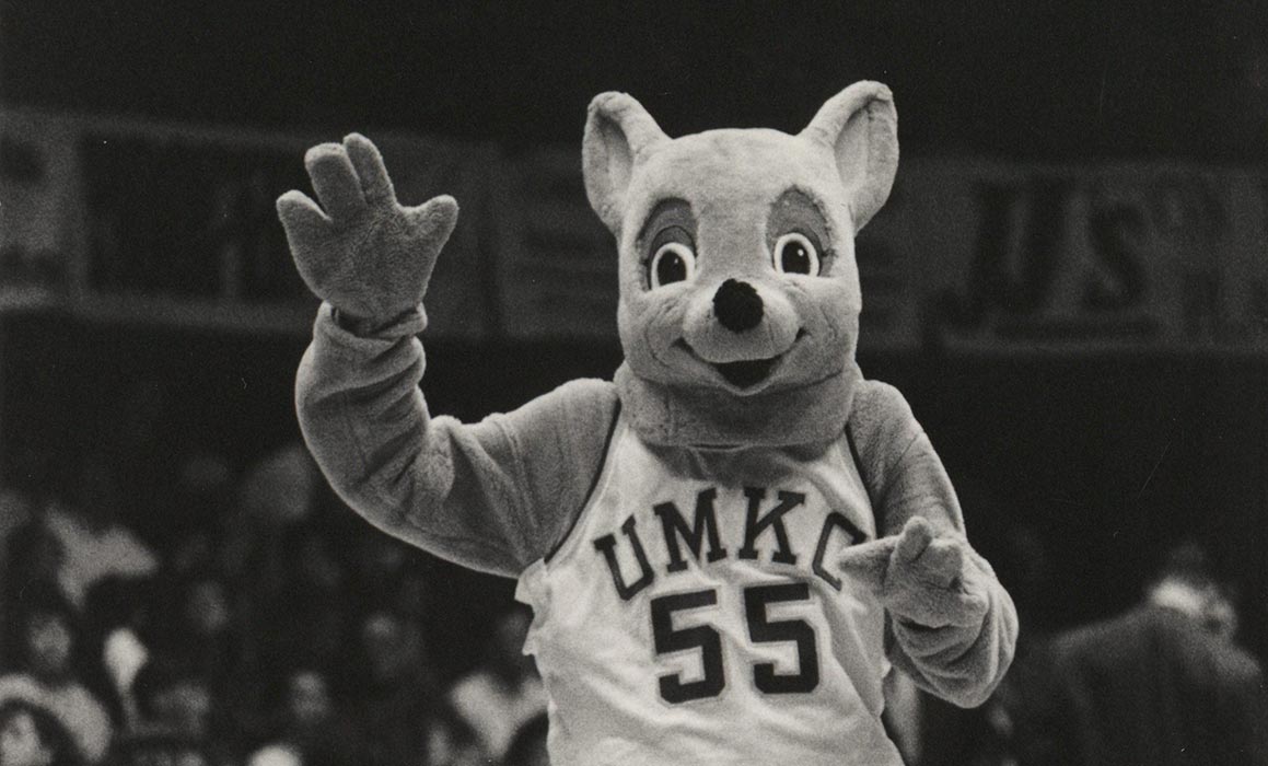 Black and white photo of UMKC mascot Kasey Roo in basketball jersey waving with people in stands behind him