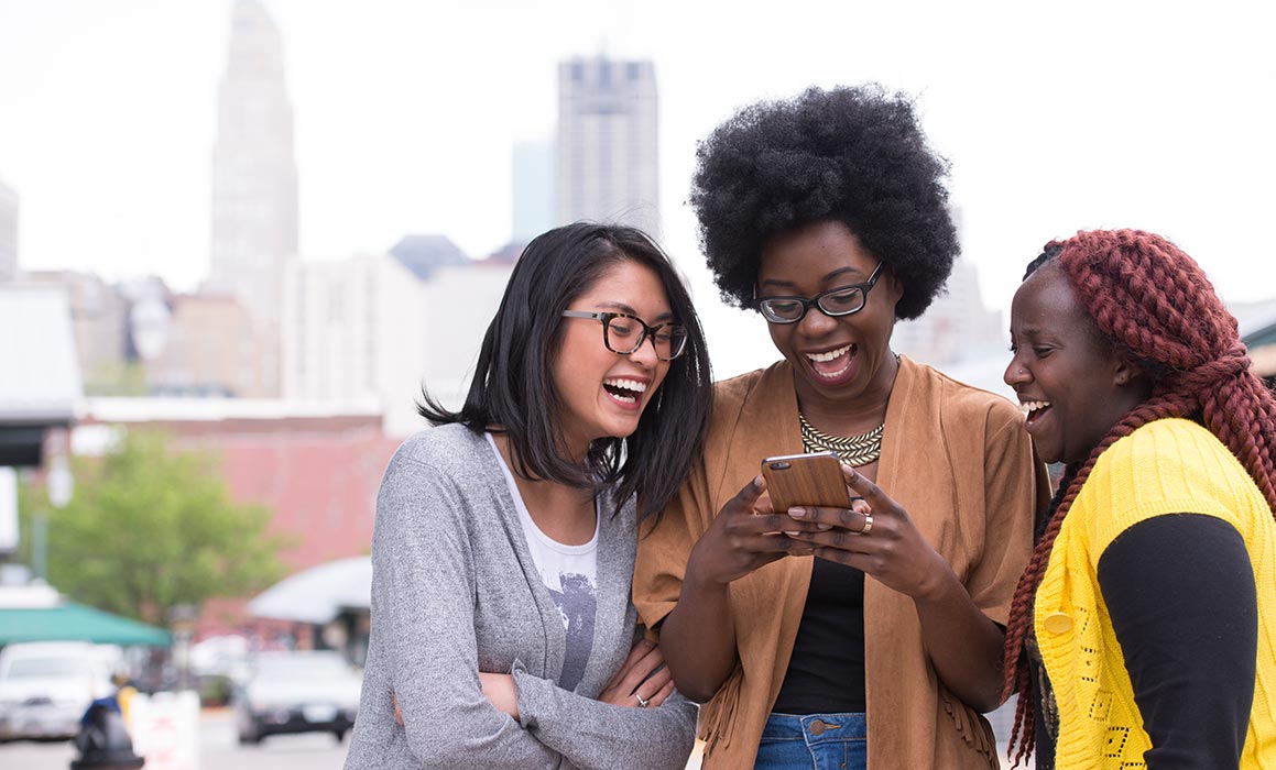 Three students presenting as women of color stand together smiling at phone with Kansas City skyline in background