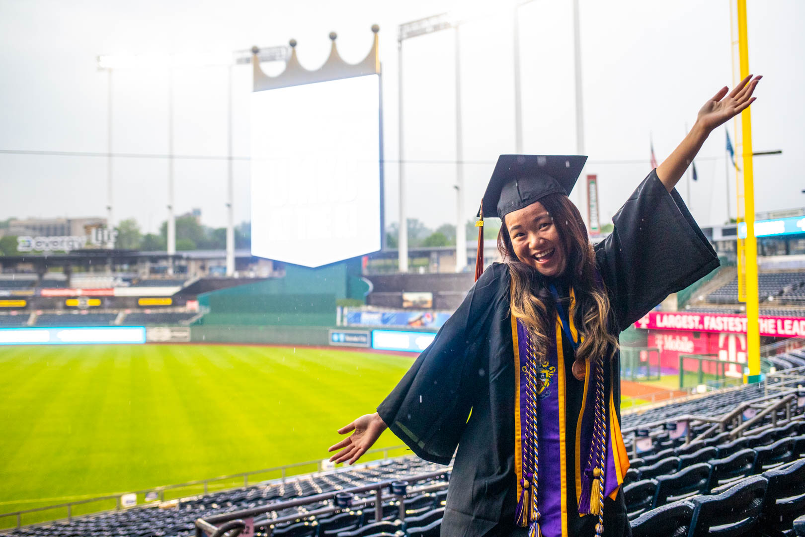 a graduate wearing a cap and gown poses in the seat at Kauffman Stadium for the UMKC commencement ceremony