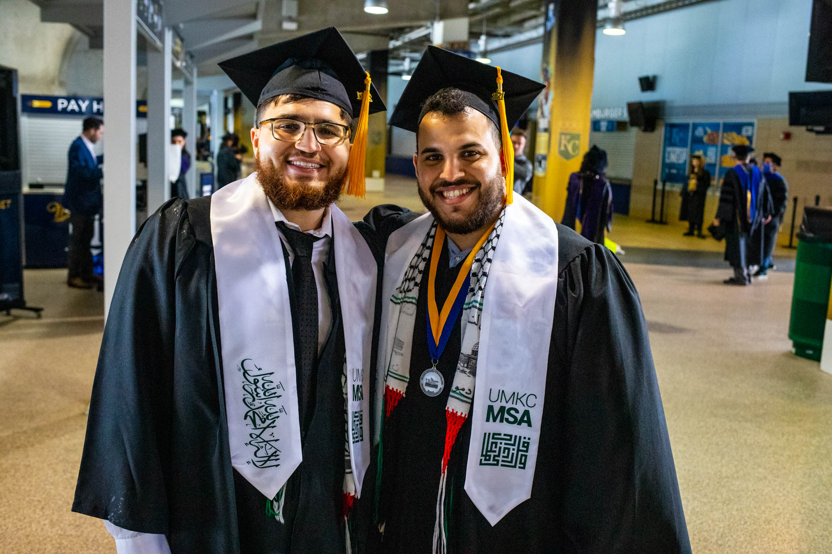 two graduates pose by each other at the commencement at Kauffman Stadium