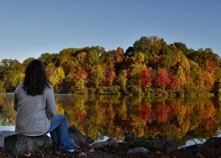 woman looking across lake at autum trees which are reflecting in the water