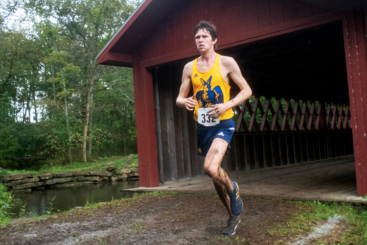 A male UMKC cross-country runner is covered in mud and running near a red covered bridge.