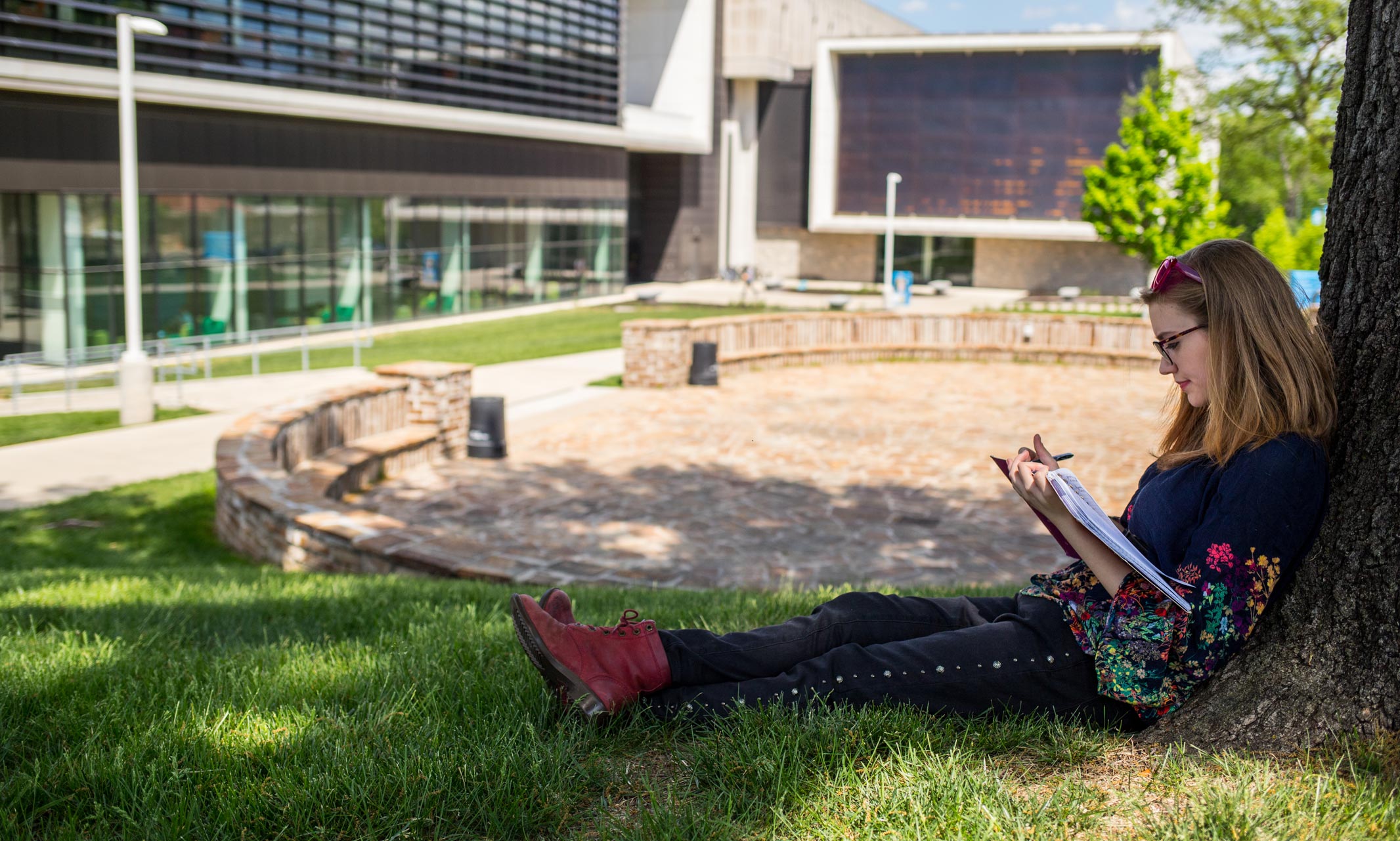Girl sits in grass next to a tree and reads a book with the UMKC Miller Nichols Library in the background
