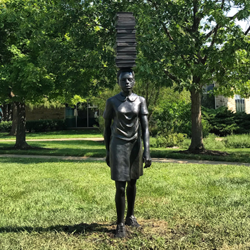 The sculpture by Flávio Cerqueira, titled “Any Word Except Wait” features a female with a stack of books on her head taking a step forward 