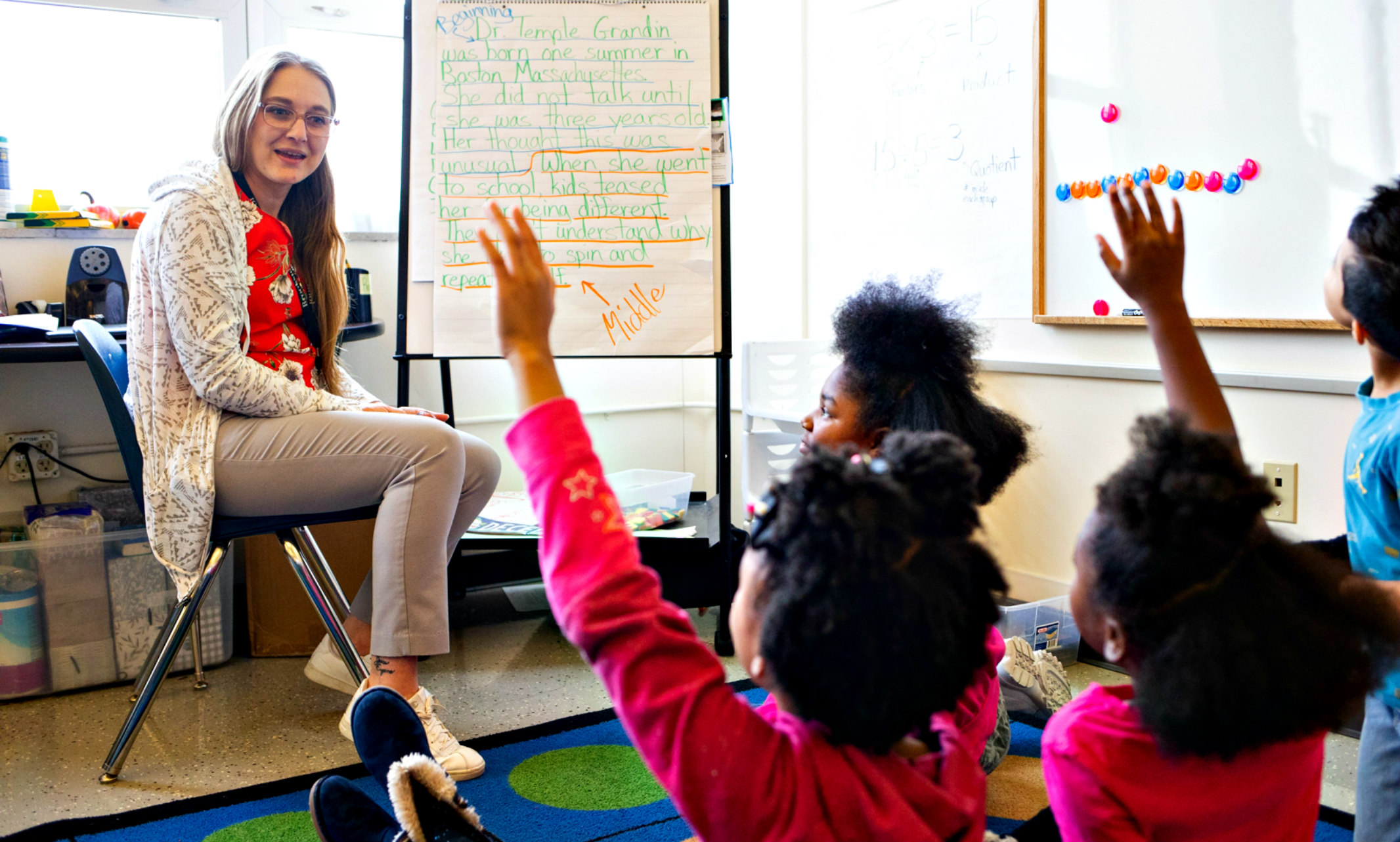 teacher sitting at front of diverse classroom with children raising hands
