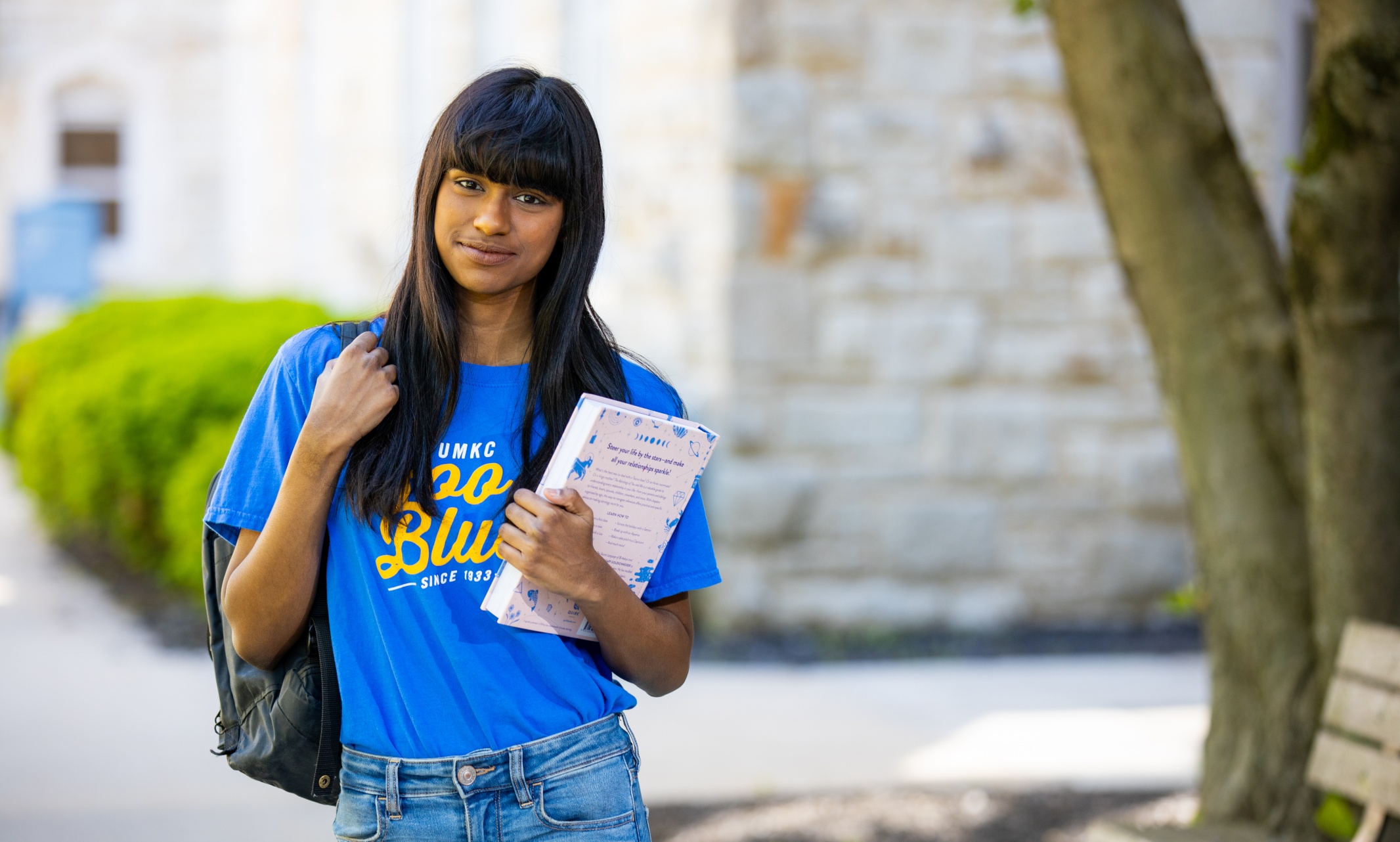 Krithika with her bookbag and a book outside on campus