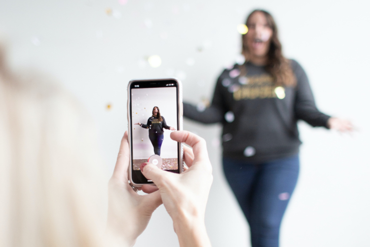 one girl takes a phone photo of another girl in the background surrounded by falling confetti