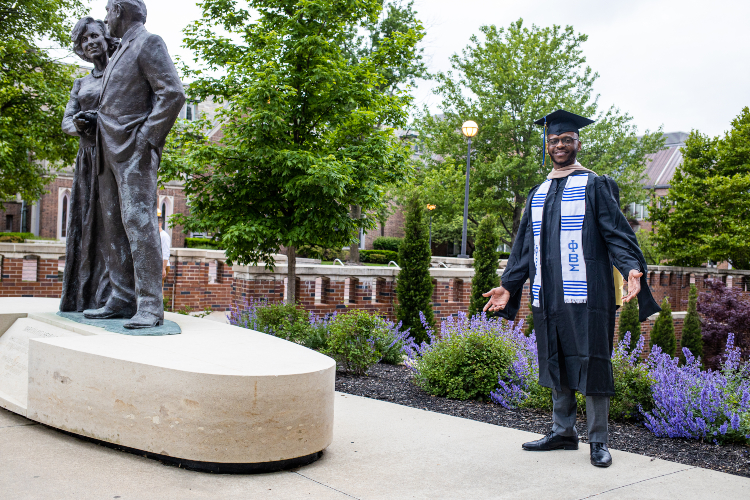 student in grad cap and gown next to statue of Henry and Marion Bloch at UMKC