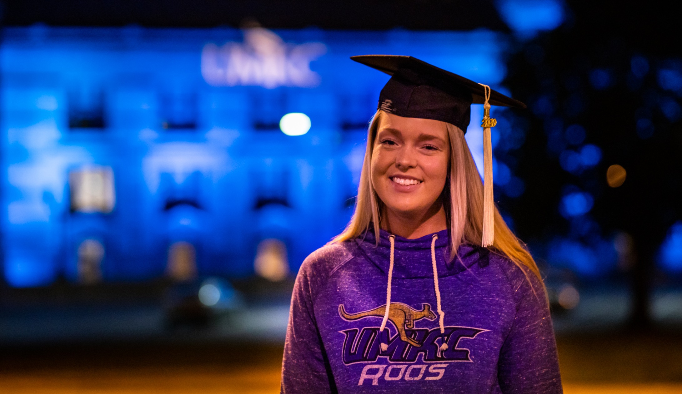 UMKC senior with grad cap stands in front of Union Station lit up in blue and gold