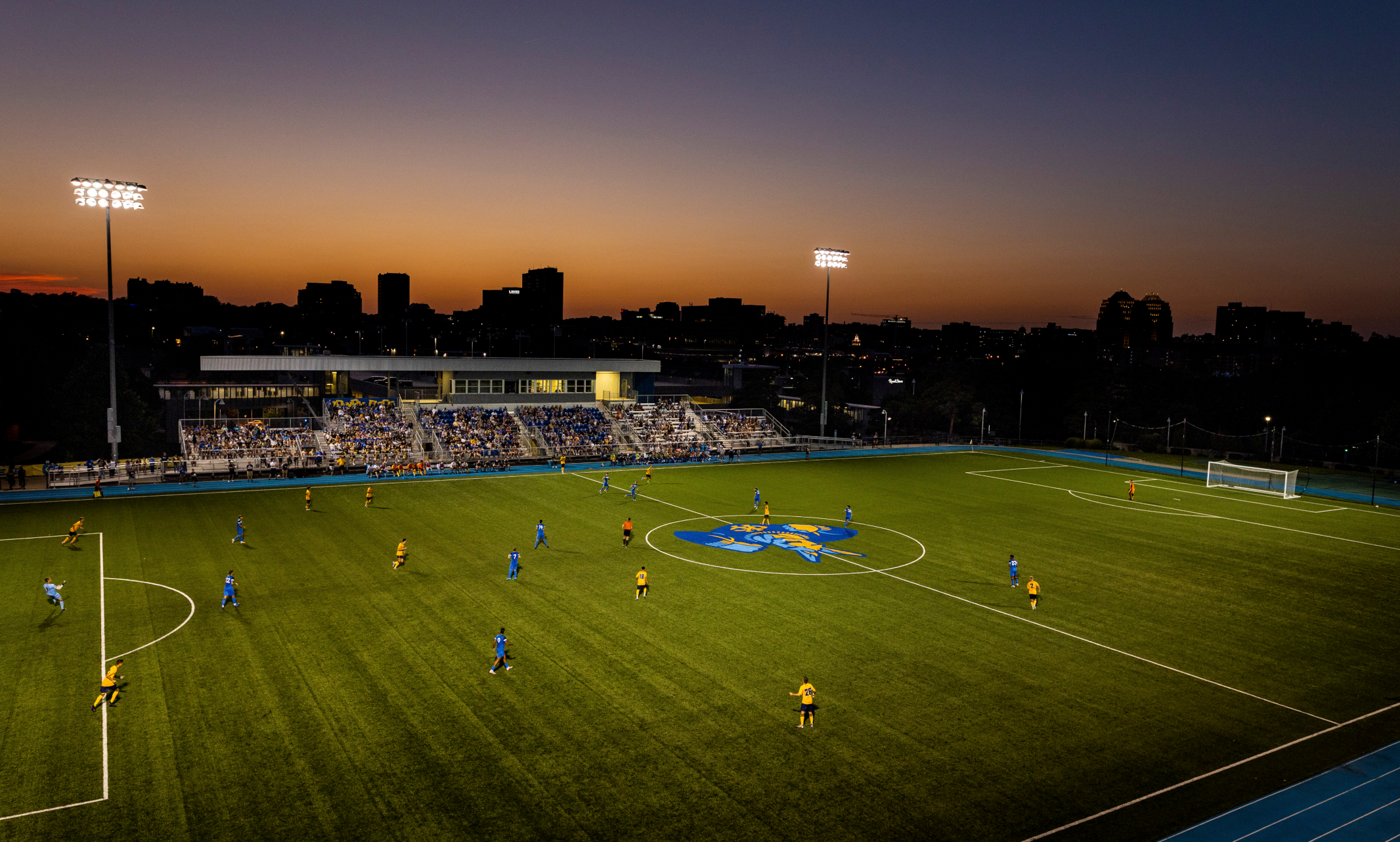 Soccer team plays at Durwood stadium at sunset with city skyline in silhouette