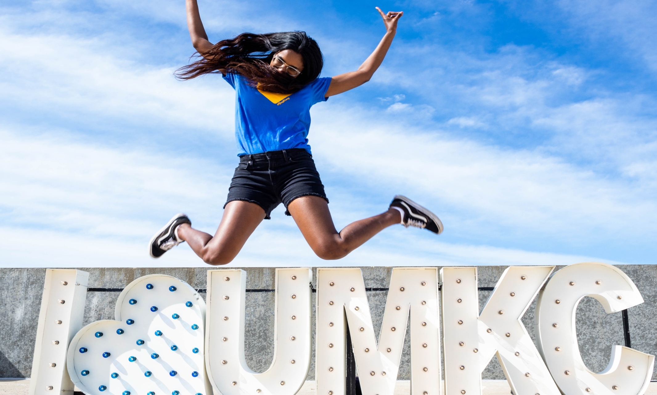 A woman in a blue shirt is jumping in front of a large light-up sign that reads 'I <3 UMKC'