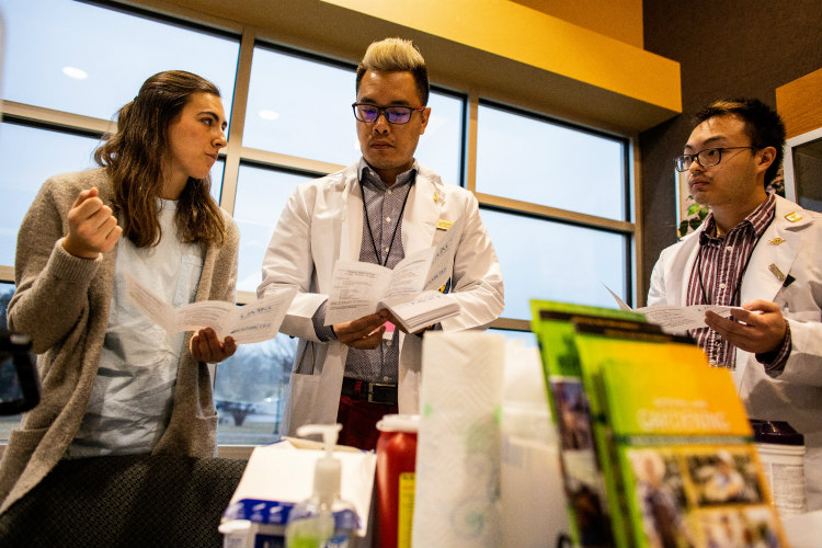 A female talks to two males; health brochures and supplies are in the foreground.