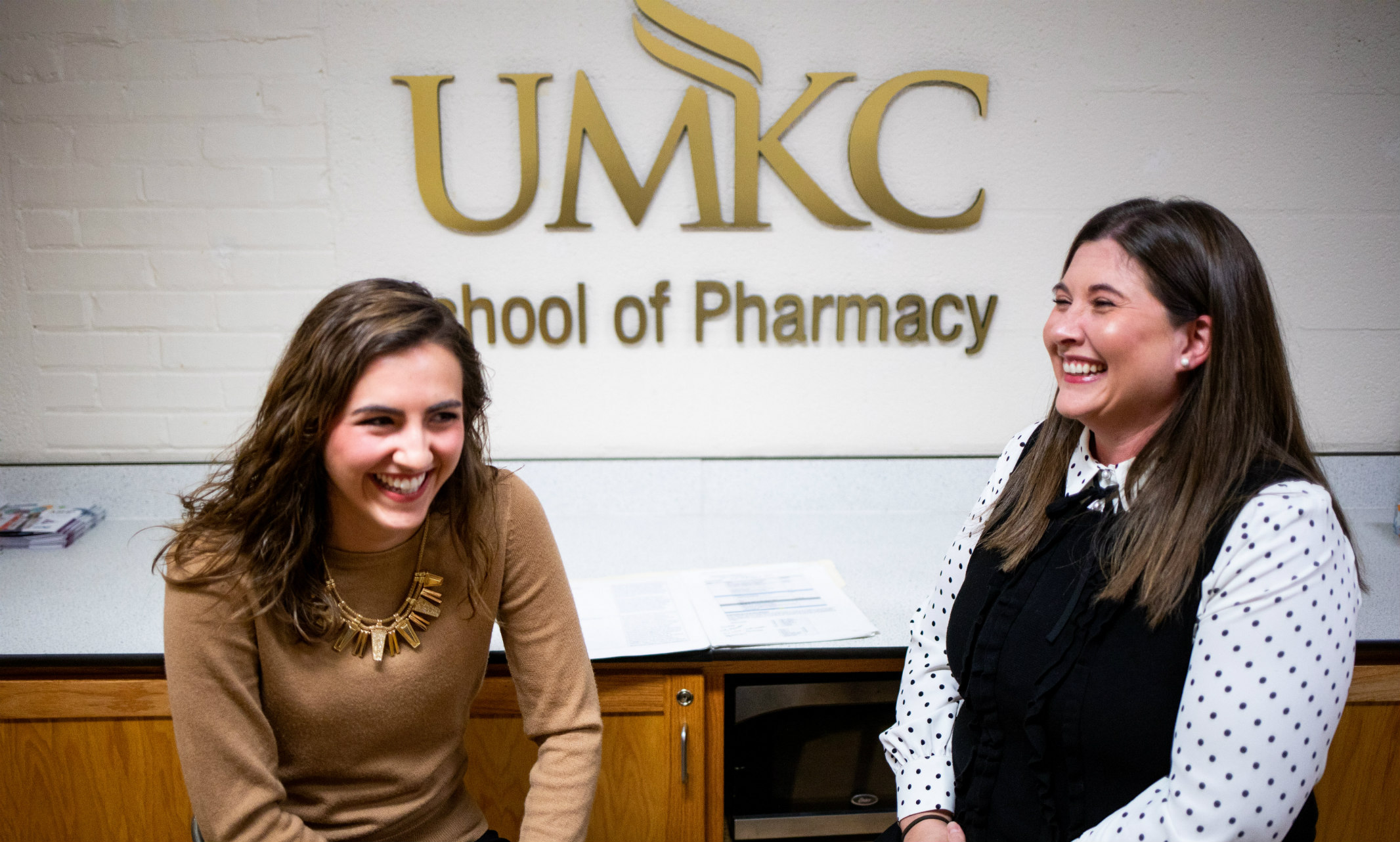 Two women sit in front of a UMKC School of Pharmacy wall sign