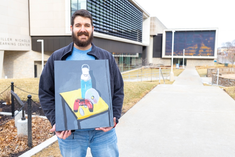 Eric stands outside Miller Nichols Library holding one of his paintings, which depicts a video game controller and military dog tags