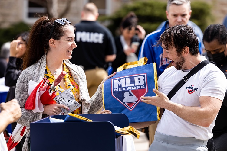 An MLB representative hands an MLB-branded tote bag and bags of chips to a person. Both are smiling.