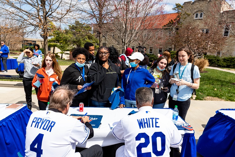 A group of students stand in front of a table to get autographs from former players Greg Pryor and Frank White, who are sitting at the table in jerseys.