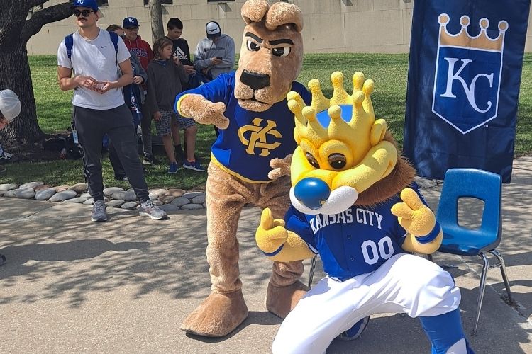 Mascots Sluggerrr and KC Roo pose together outside, in front of a Royals banner