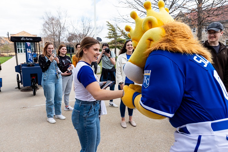 Sluggerrr holds a students hand; the student holds her arm out and laughs at Sluggerrr. Other students are in the background laughing and taking pictures.