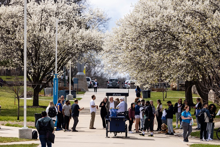 A crowd of people stands and walks on a large sidewalk, an MLB cart is in the middle. Flowering trees line both sides of the sidewalk against a blue cloudy sky.