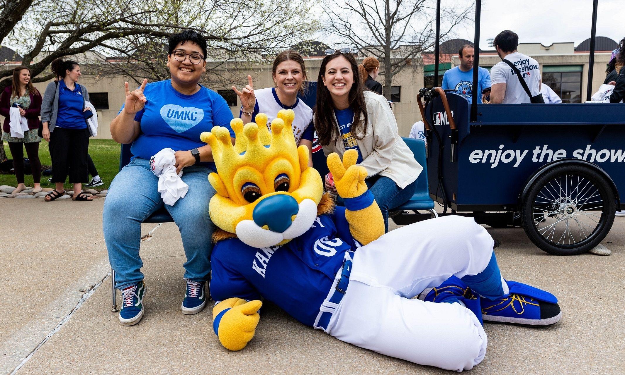 Three students sit in chairs outside, holding "Roo Up" symobls and smiling. Sluggerrr mascot lays on the ground in front of them looking at the camera.