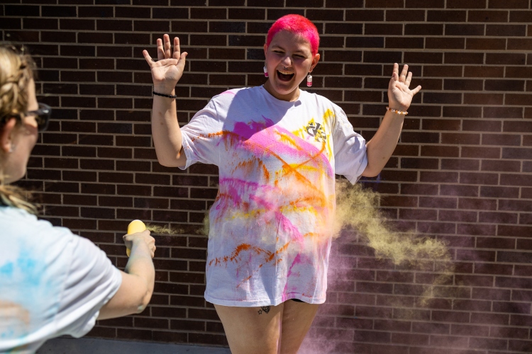 A student stands with arms out as people toss color powder on their shirt