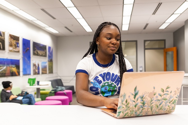 Symone Franks works on her laptop in a large, brightly-lit room. A student, out of focus, sits in a chair in another part of the room.