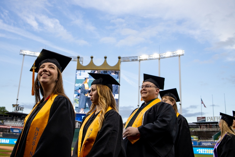 Three students stand in cap and gowns with Kauffman Stadium's Crown Vision screen in the background