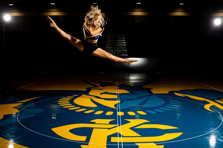 A dancer leaps in the air with her legs in a splits position. Below her is the Roo that is painted on the floor of the Swinney basketball court.