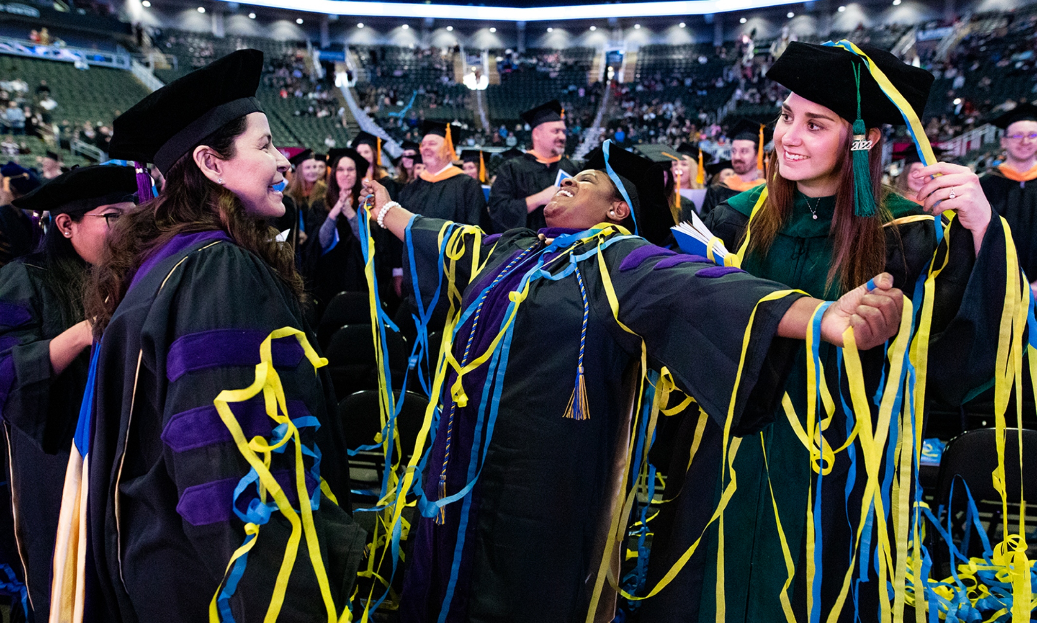 A graduate stands with her arms outstretched covered in blue and gold streamers, smiling with two other graduates looking at her and smiling