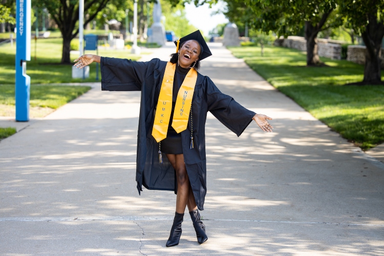 A graduate stands joyous on university walkway with one foot over the other, arms stretched out and head tilted, smiling