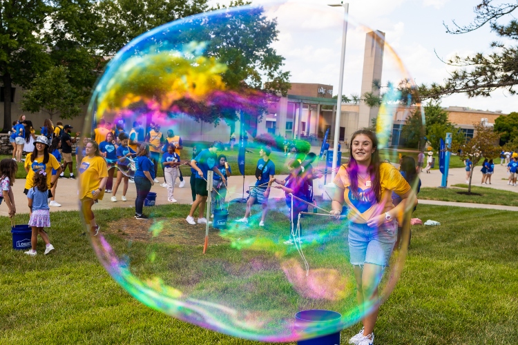 A translucent bubble is in the foreground with a student behind it, holding a bubble wand and smiling on a sunny university walkway