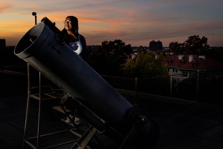 A student stands behind a large telescope at Wako Observatory at dusk