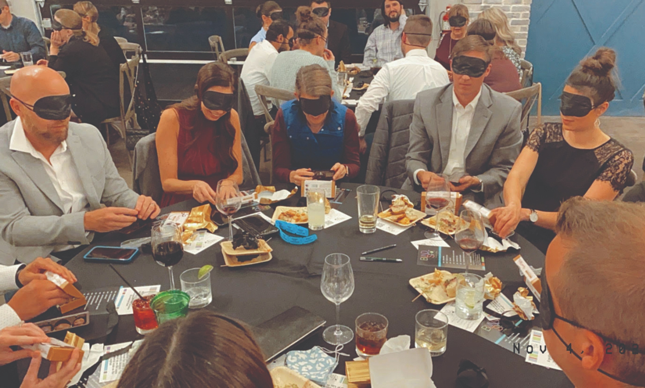 A group of people sit at a table having a meal while wearing blindfolds 