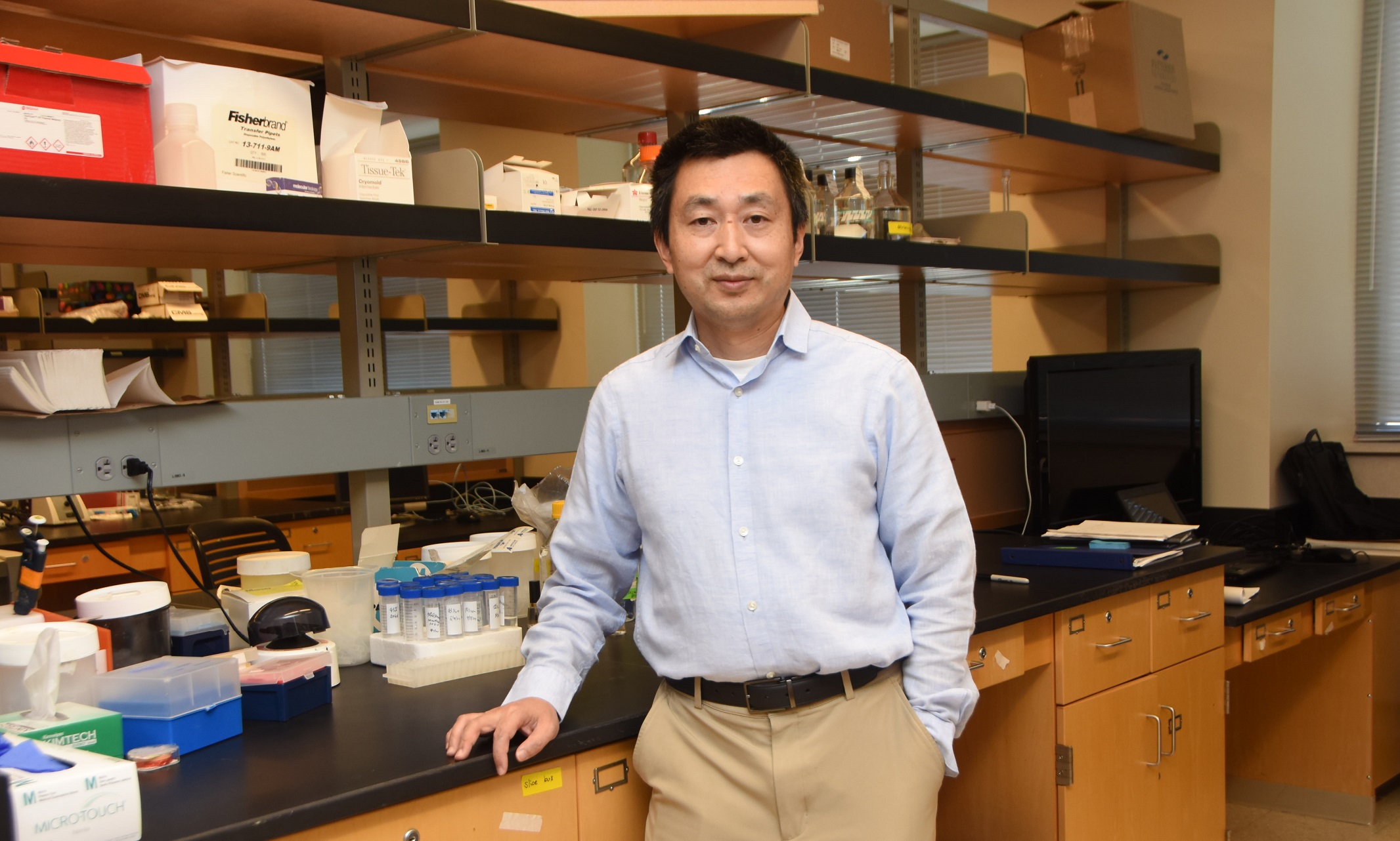 Xiangming Zha stands in his lab and smiles at the camera while leaning against a counter filled with equipment.