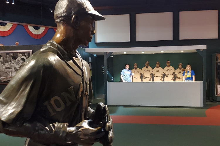 Hunter and Mariah stand in a dugout with a statue of a pitcher in the foreground